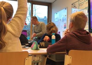 Foto van leerlingen in een klas; de leraar zit op zijn bureau en een meisje steekt haar hand op